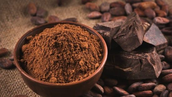 A bowl of cocoa powder next to pieces of chocolate and cocoa beans