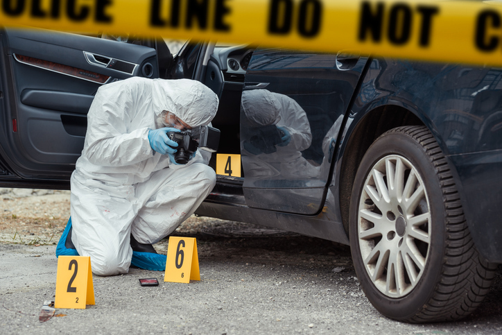 A forensic specialist suited up at crime scene next to car examining and preparing evidence through ASTM E860 methods.