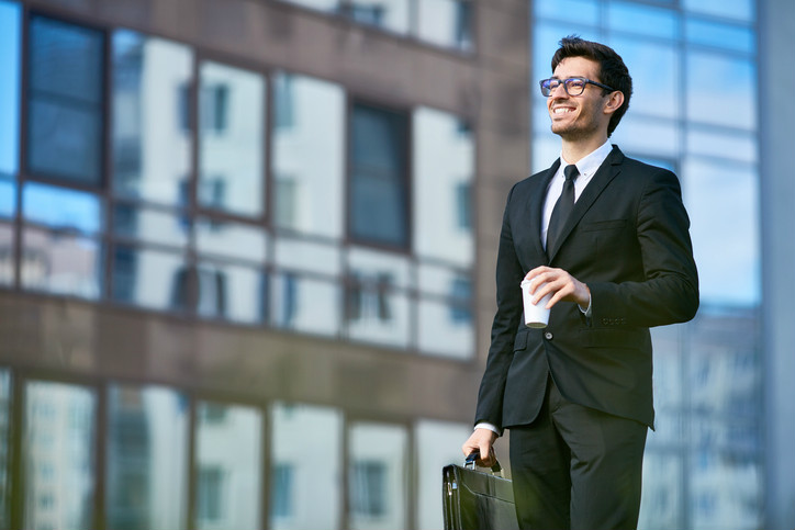 A man in a suit holding a coffee cup and a briefcase is smiling off into the distance which is symbolic for ISO 9004