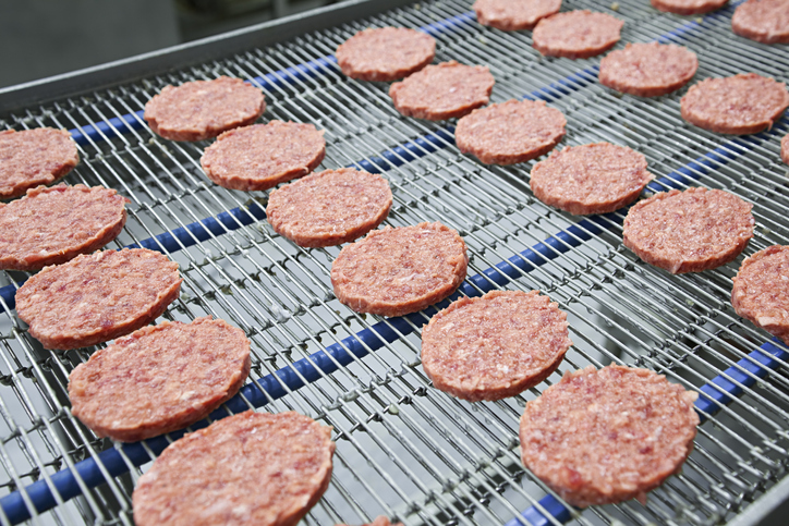 Burger patties sliding across NSF/ANSI/3A 14159-1-2019 meat processing equipment in a food plant.