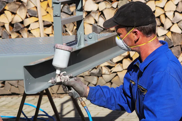 Worker in mask spraying steel with ISO 12944 corrosion protection paint.