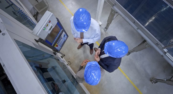 Three managers in blue hardhats abiding to ANSI B11 standards while looking at a safe machine in factory.
