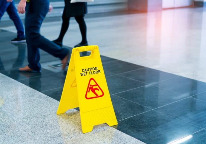 A bright yellow wet floor sign set before walking people on a walkway tested for NFSI B101.1-2020 wet SCOF.