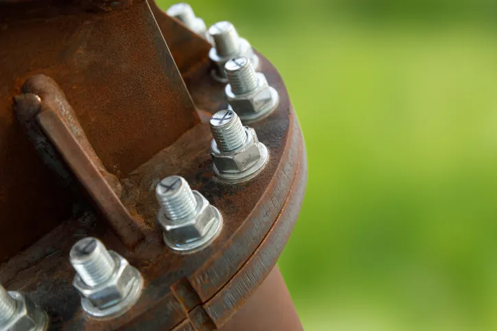 Chrome screws poking out of rusty petroleum piping system with ASME A13.1-2020 marking.