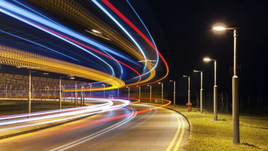 Cars pass by in streaming colors beside roadway lighting marked to ANSI C136.15-2020 luminaire identification.