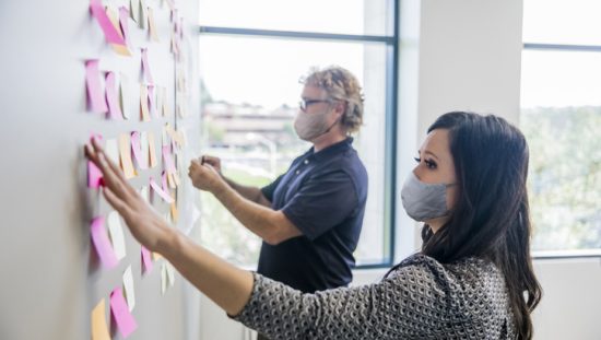 Two project managers wearing face masks and organizing tasks in accordance with ISO 21502:2020 guidance.