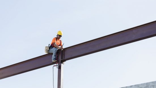 Worker sitting on girder and held safe by ANSI/ASSP Z359.11-2021 full body harness in the sky.