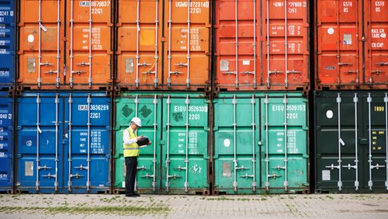 Manager walking beside colorful shipping containers and checking on supply chain security through SCS 9001.