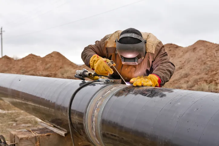 Worker Welding Pipeline Worker covered in PPE in the plains welding a black pipeline with proper ASME B31Q-2021 pipeline personnel qualifications.
